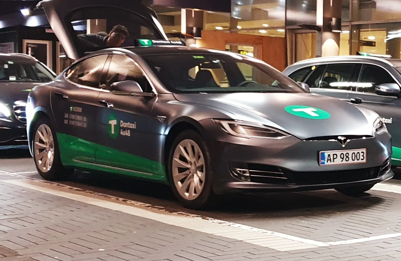 Grå Tesla Model S med grøn taxafolie holder med åben frunk foran en oplyst lufthavnsterminal om aftenen