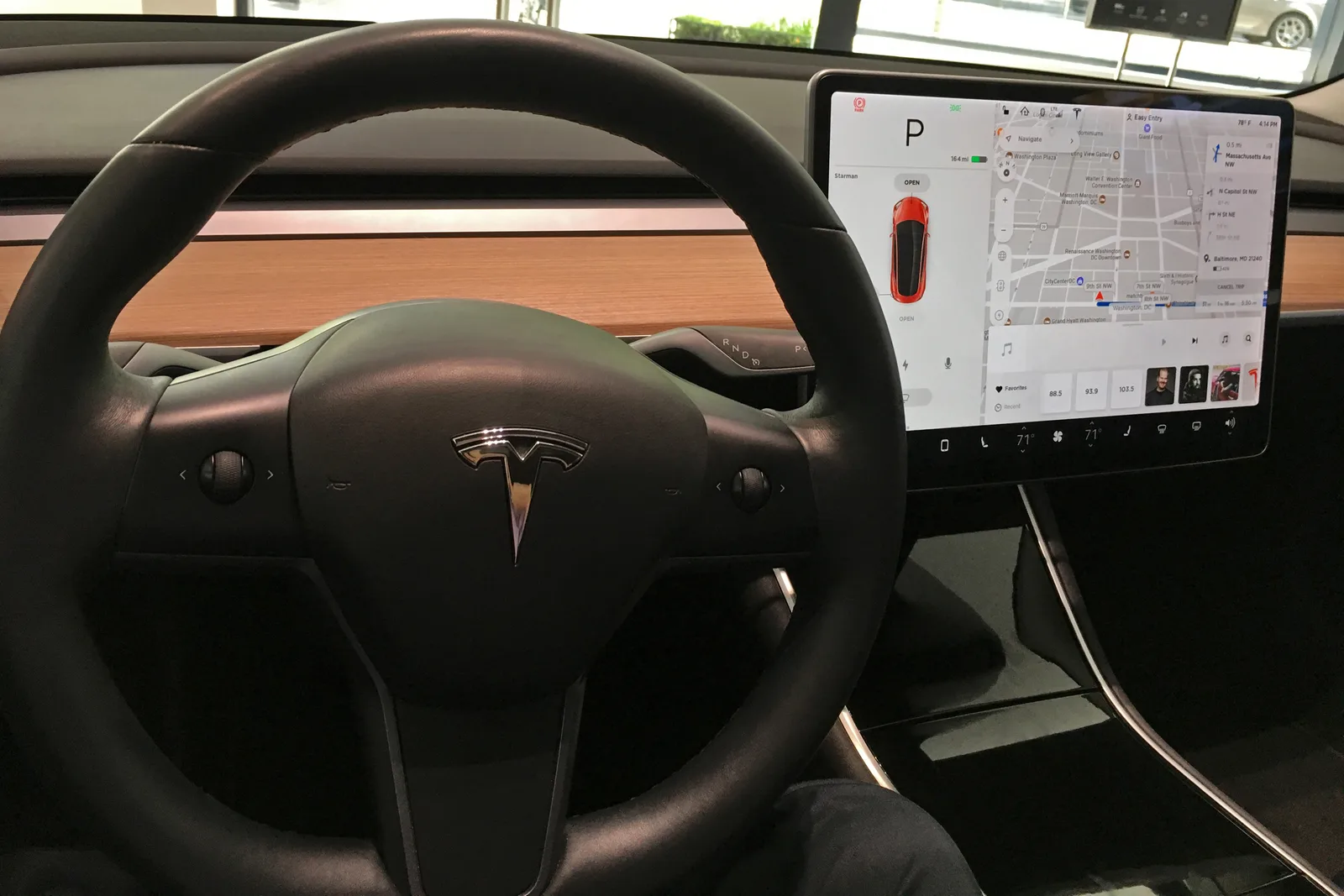Førerpladsen i en Model 3 fra 2018 med sort rat, to rullehjul, trægreb hen over instrumentbrættet og den tændte midterskærm