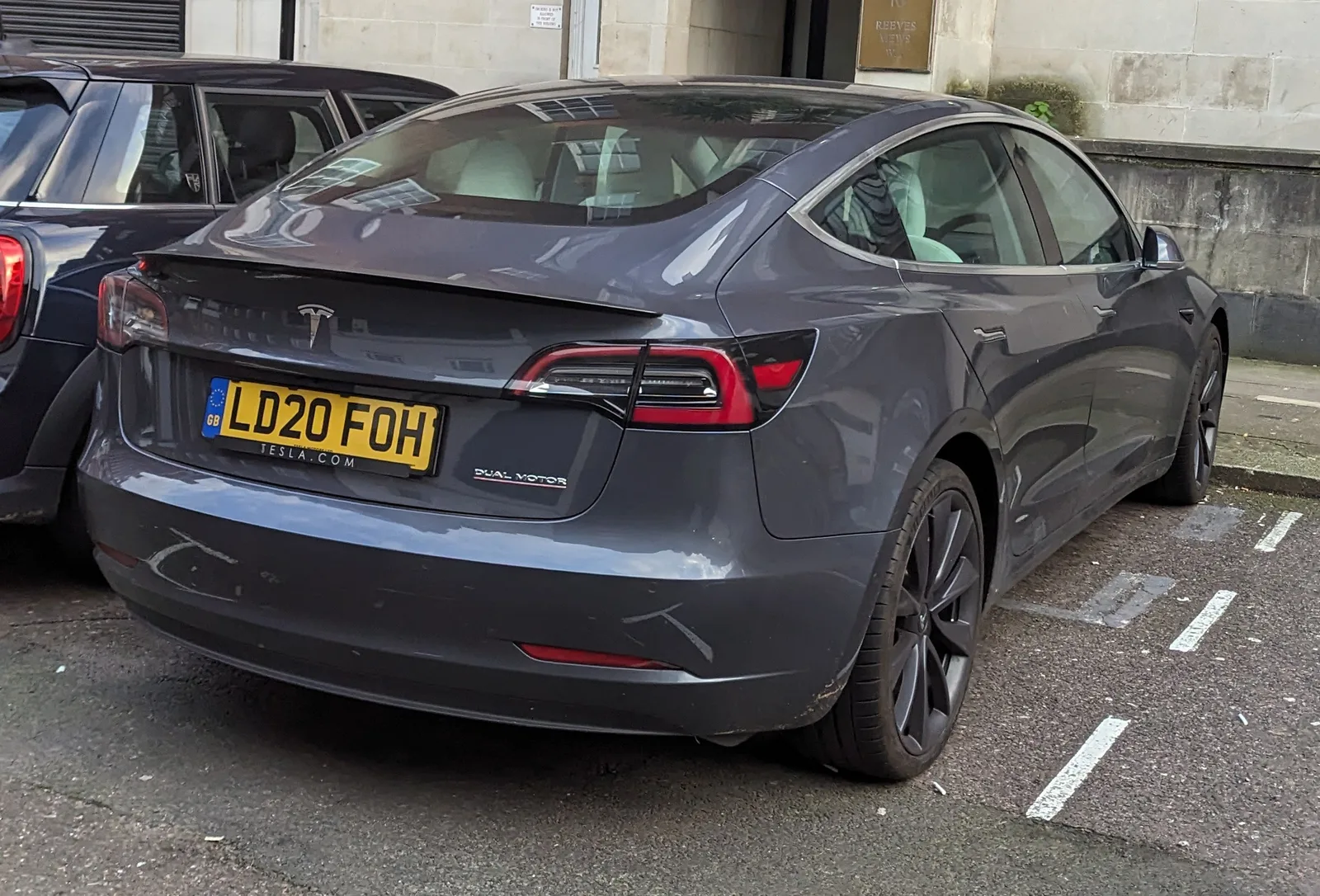 En grå Tesla Model 3 fra 2020 set skråt bagfra, parkeret ved en sandstensfacade i en bygade