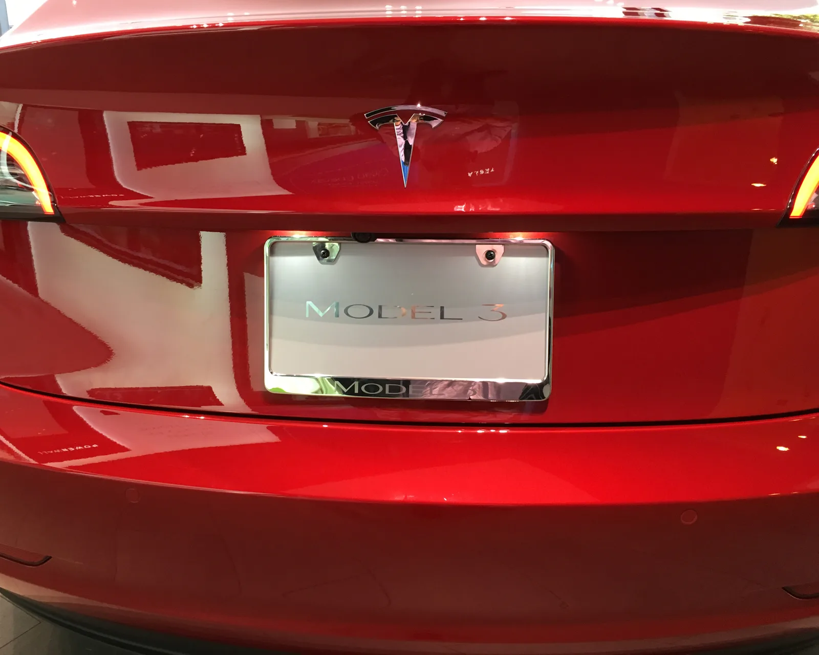 Bagenden af en rød Tesla Model 3 tæt på, med Tesla-mærket og en skilteramme, hvor der står MODEL 3 i stedet for en nummerplade