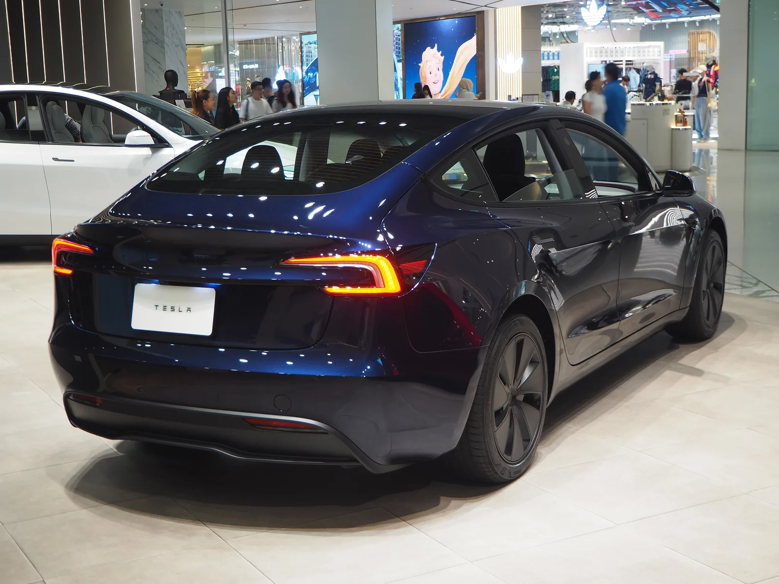 Mørkeblå Tesla Model 3 set skråt bagfra i en Tesla-butik i et indkøbscenter, med en hvid Model Y ved siden af og butikker i baggrunden
