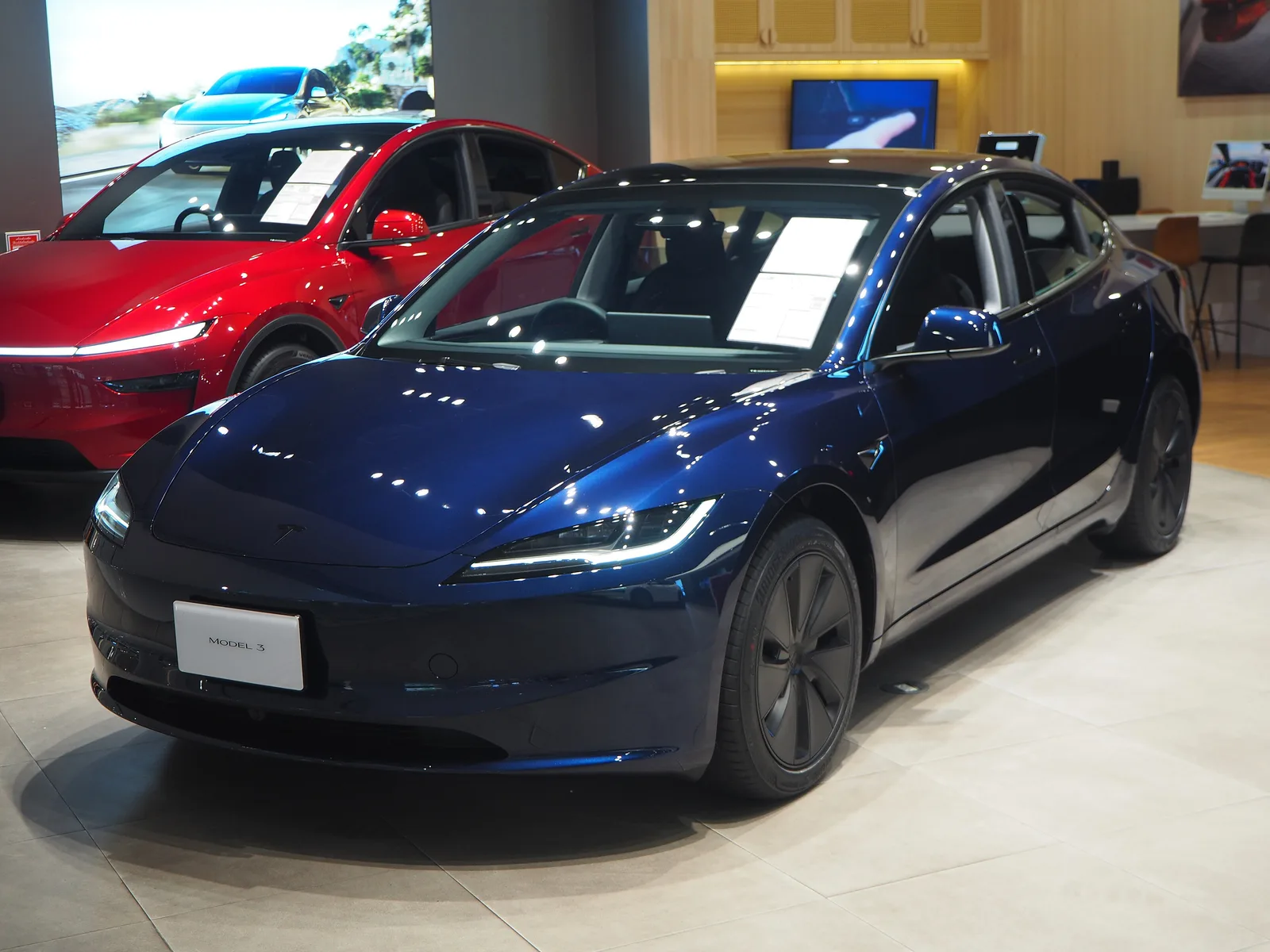 Mørkeblå Tesla Model 3 set skråt forfra i en Tesla-butik med prisseddel i forruden og en rød Model Y bag den