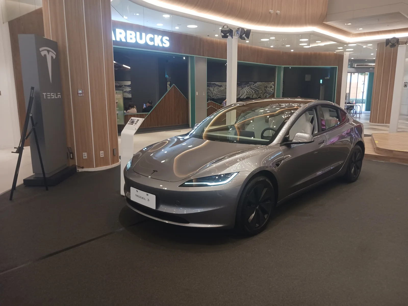 En grå Tesla Model 3 udstillet på en stand i et indkøbscenter, med en Tesla-søjle og en infoskærm ved siden af og butikker i baggrunden
