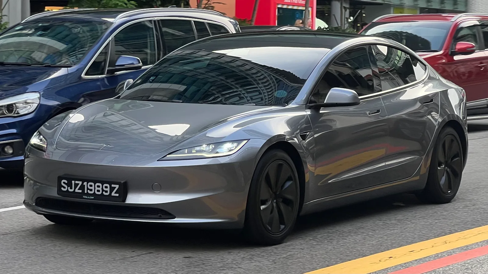 Grå Tesla Model 3 Highland set skråt forfra i tæt bytrafik med en blå og en rød bil på hver side