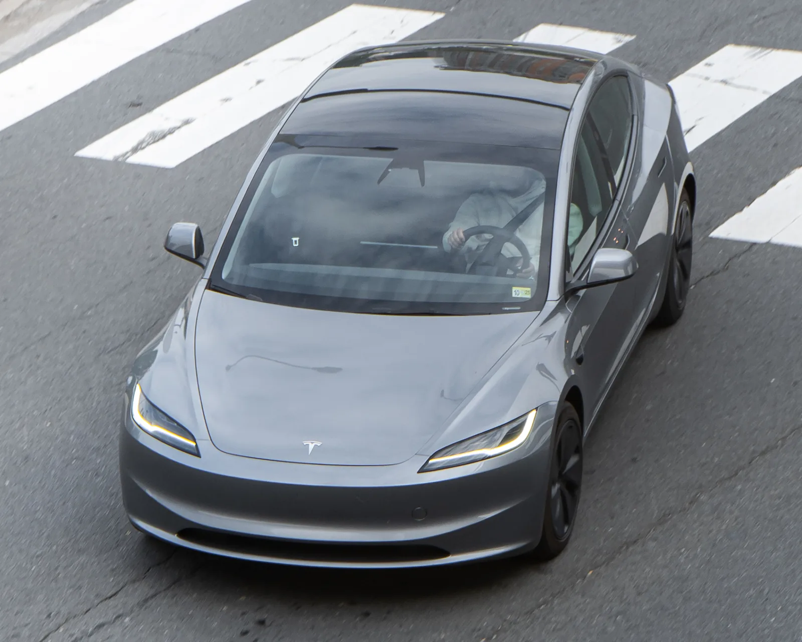 Grå Tesla Model 3 Highland set skråt oppefra under kørsel over et fodgængerfelt, hele glastaget og motorhjelmen synlig