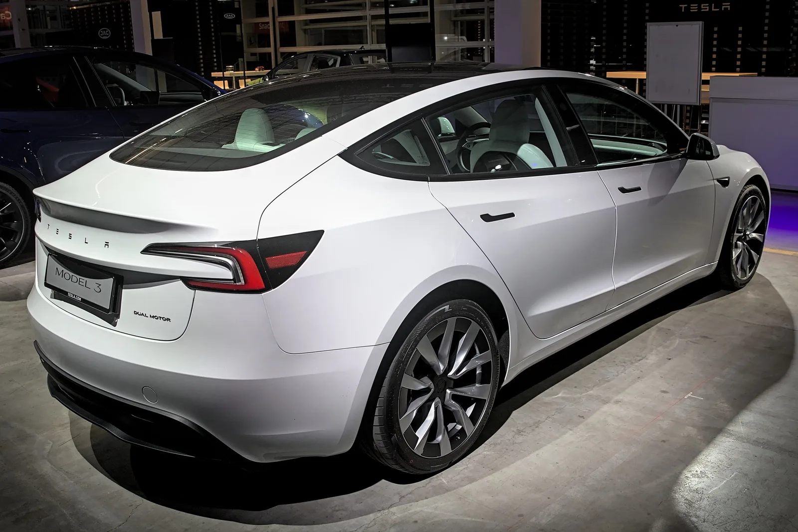 Hvid Tesla Model 3 af den nye udgave set skråt bagfra i et udstillingslokale, med Dual Motor-emblem og det lille dæksel over trækhuset i kofangeren