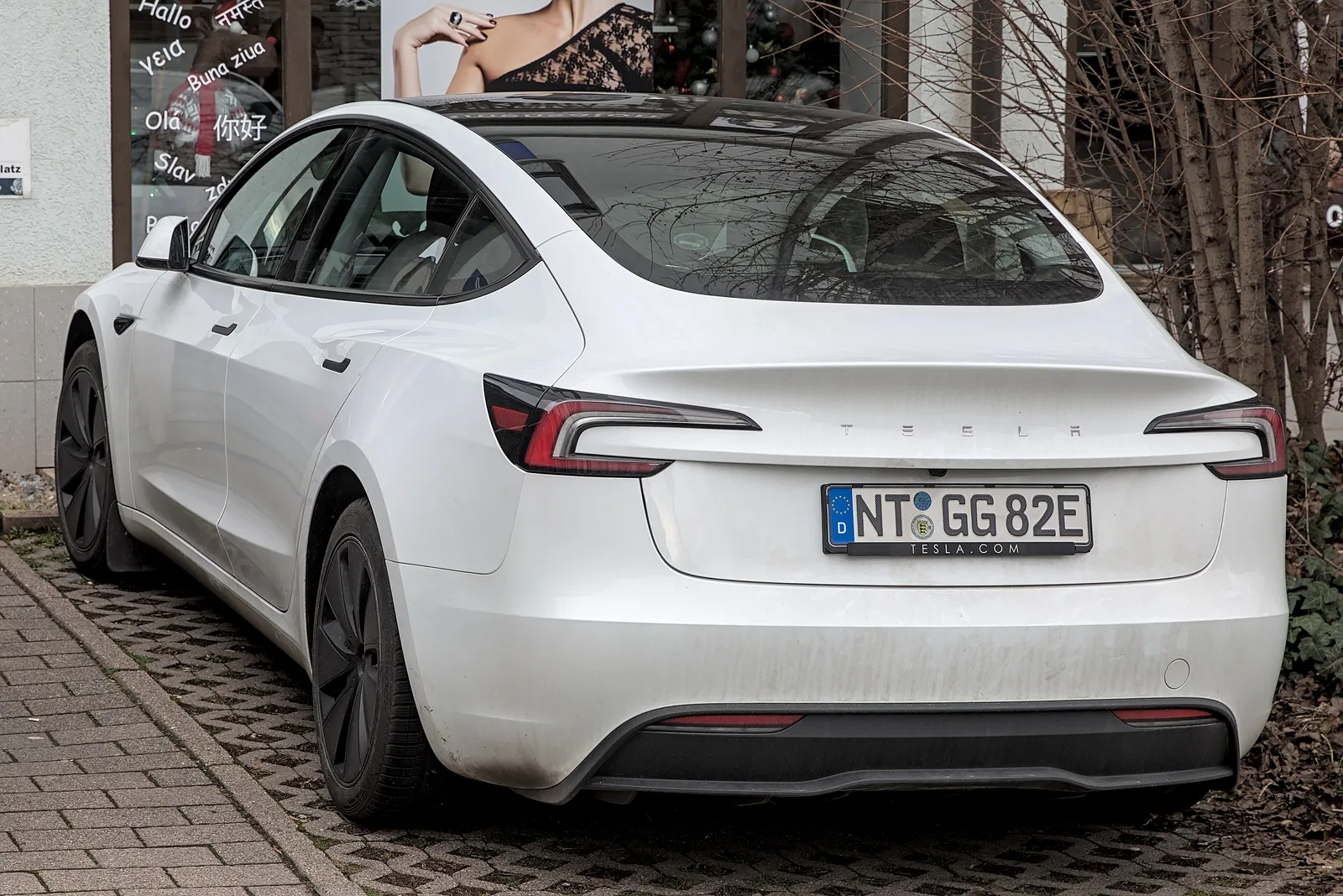 Hvid Tesla Model 3 Highland set bagfra, parkeret ved en butiksfacade med nøgne træer om vinteren