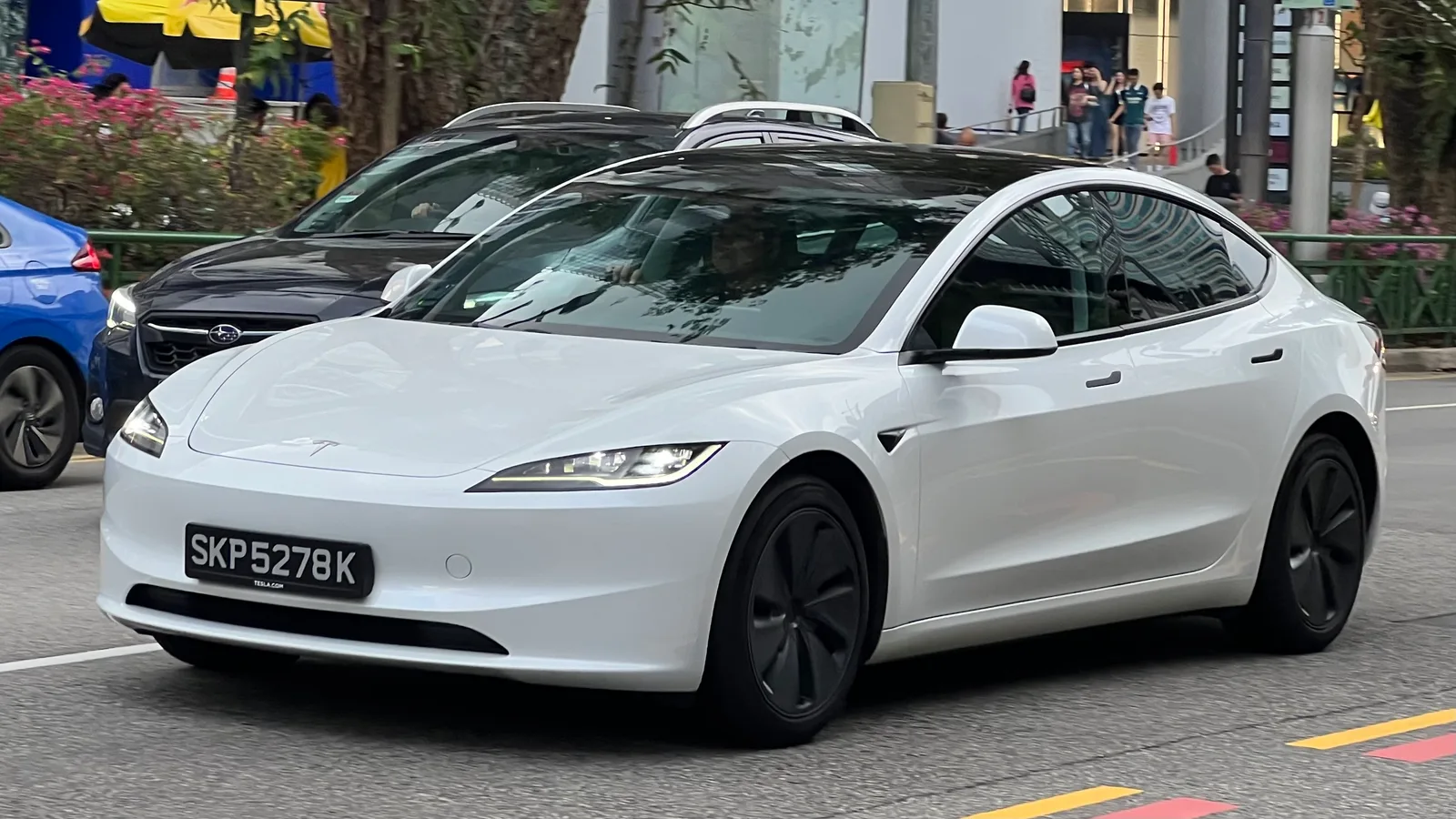 Hvid Tesla Model 3 set skråt forfra i tæt bytrafik med forlygterne tændt og fodgængere på fortovet bag