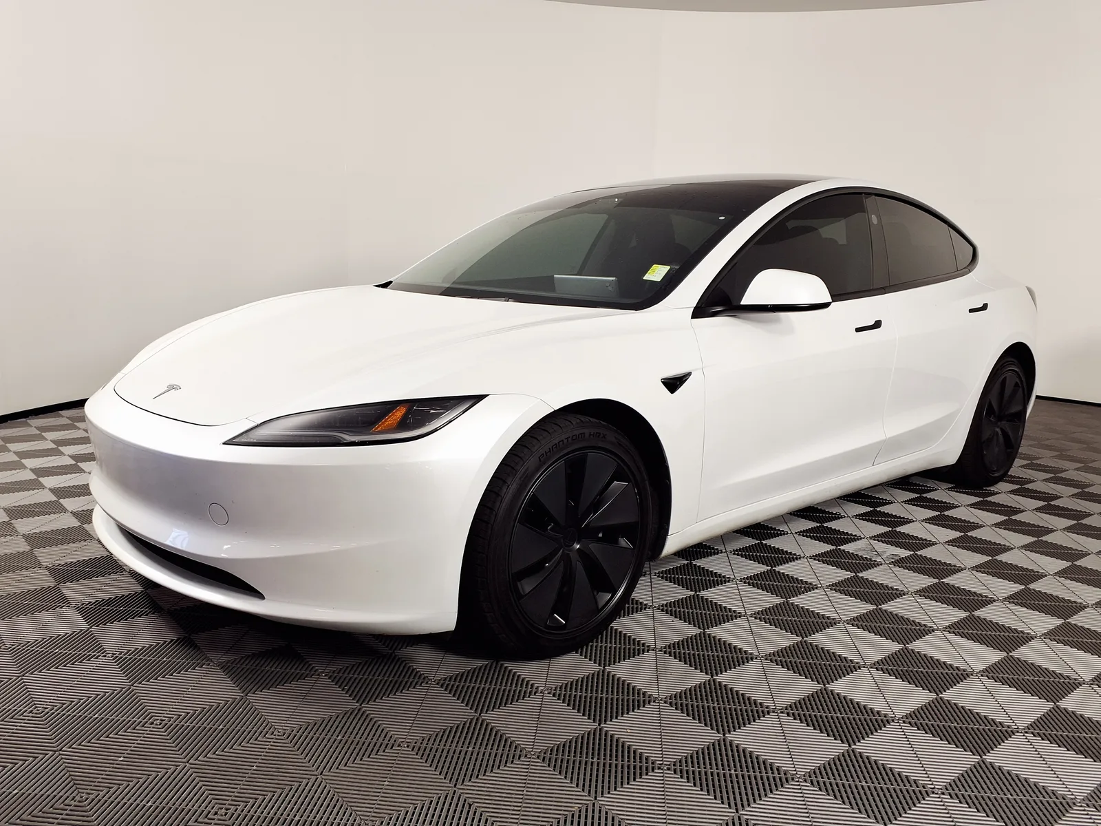 En hvid Tesla Model 3 i basisudgaven med sorte aerofælge, set skråt forfra i et fotostudie med ternet gulv