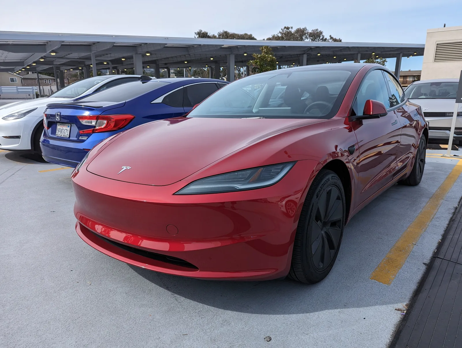 Rød Tesla Model 3 Highland set skråt forfra på en parkeringsplads under et overdækket solcelletag, med en blå Honda og en hvid Model 3 ved siden af