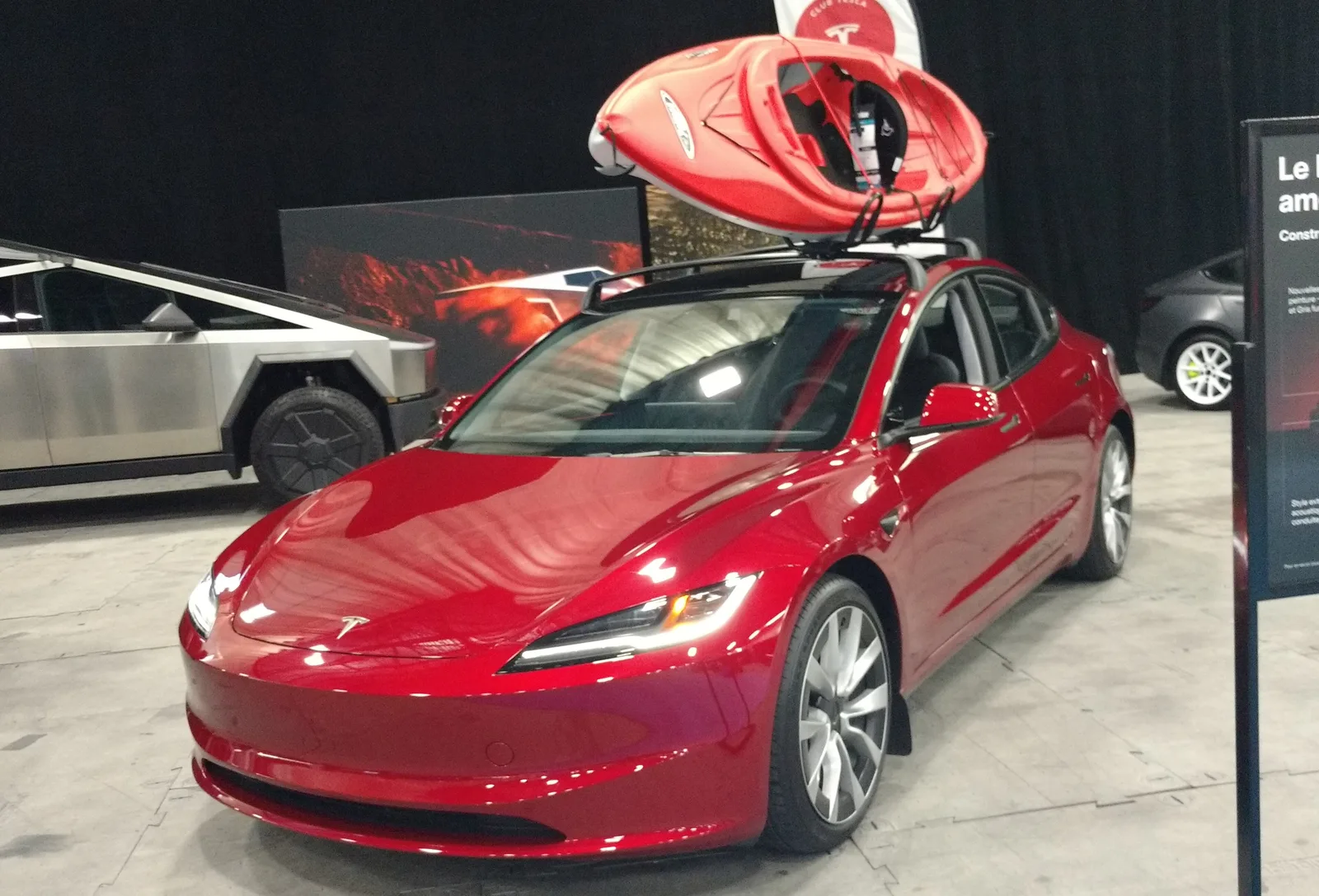 Rød Tesla Model 3 Highland med tagbøjler og en rød kajak på taget på en bilmesse, Cybertruck i baggrunden