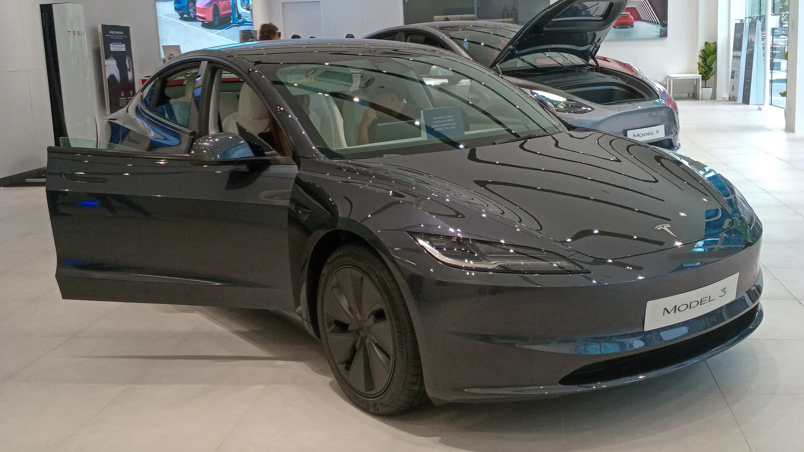 Sort Tesla Model 3 set skråt forfra i en Tesla-butik med fordøren åben, en grå Model Y med åben frunk bagved