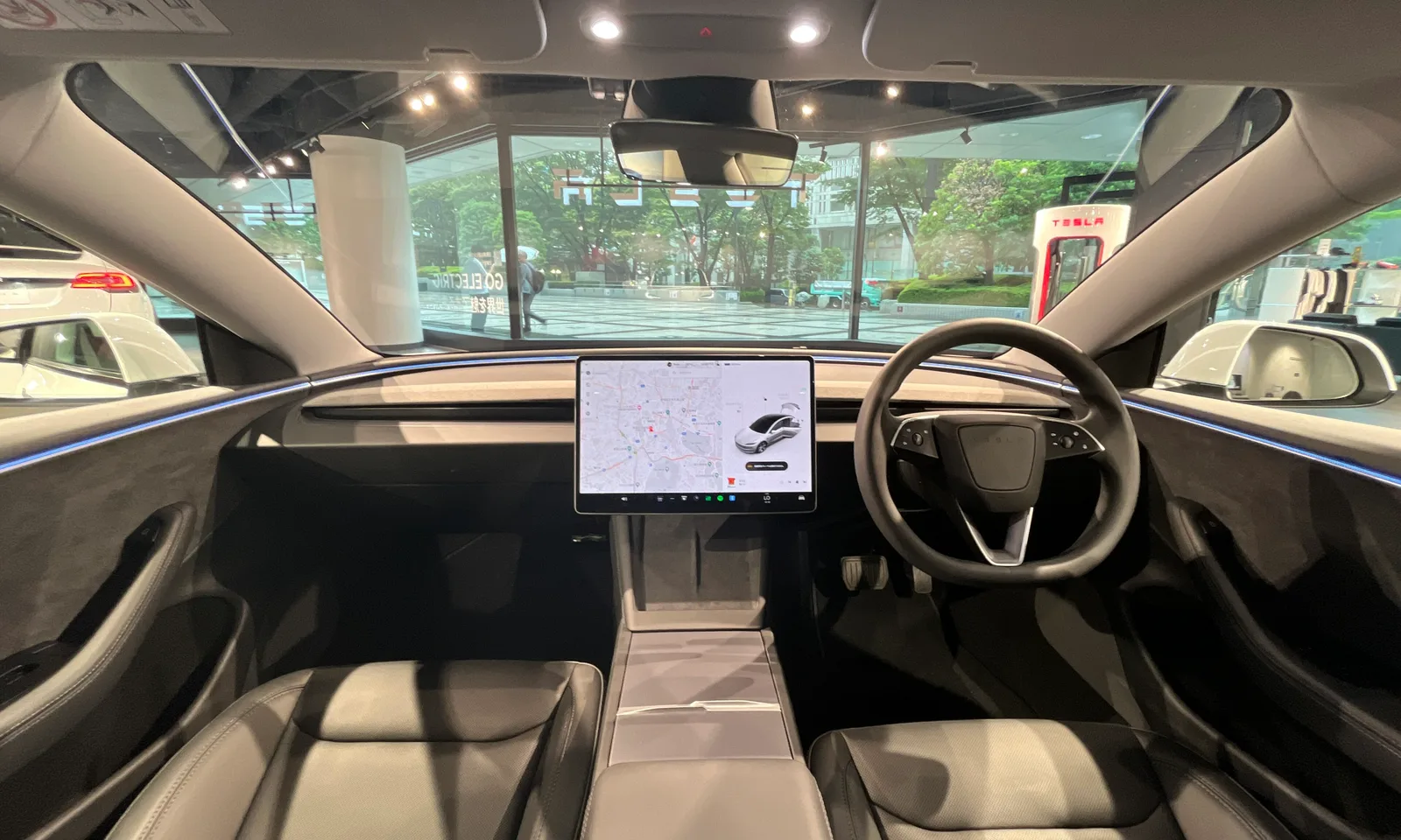 Kabinen i en Model 3 af den nye udgave set bagfra mellem forsæderne, med den tændte midterskærm, der viser et kort og bilens gengivelse, og en blå lysliste hen over instrumentbrættet