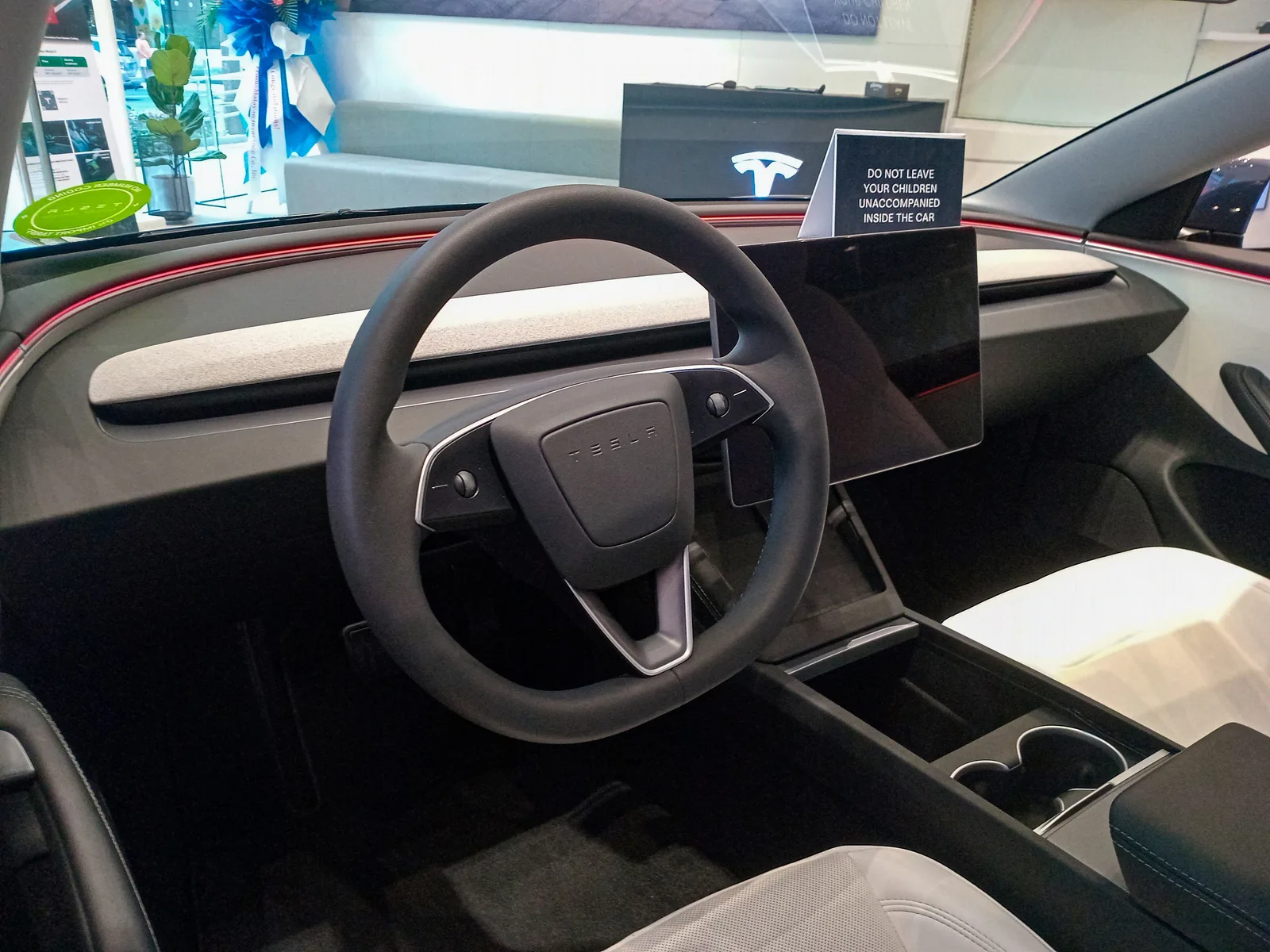 Førerpladsen i en Tesla Model 3 med rat, midterskærm og lyst instrumentbræt, set fra passagersiden i en udstillingssal