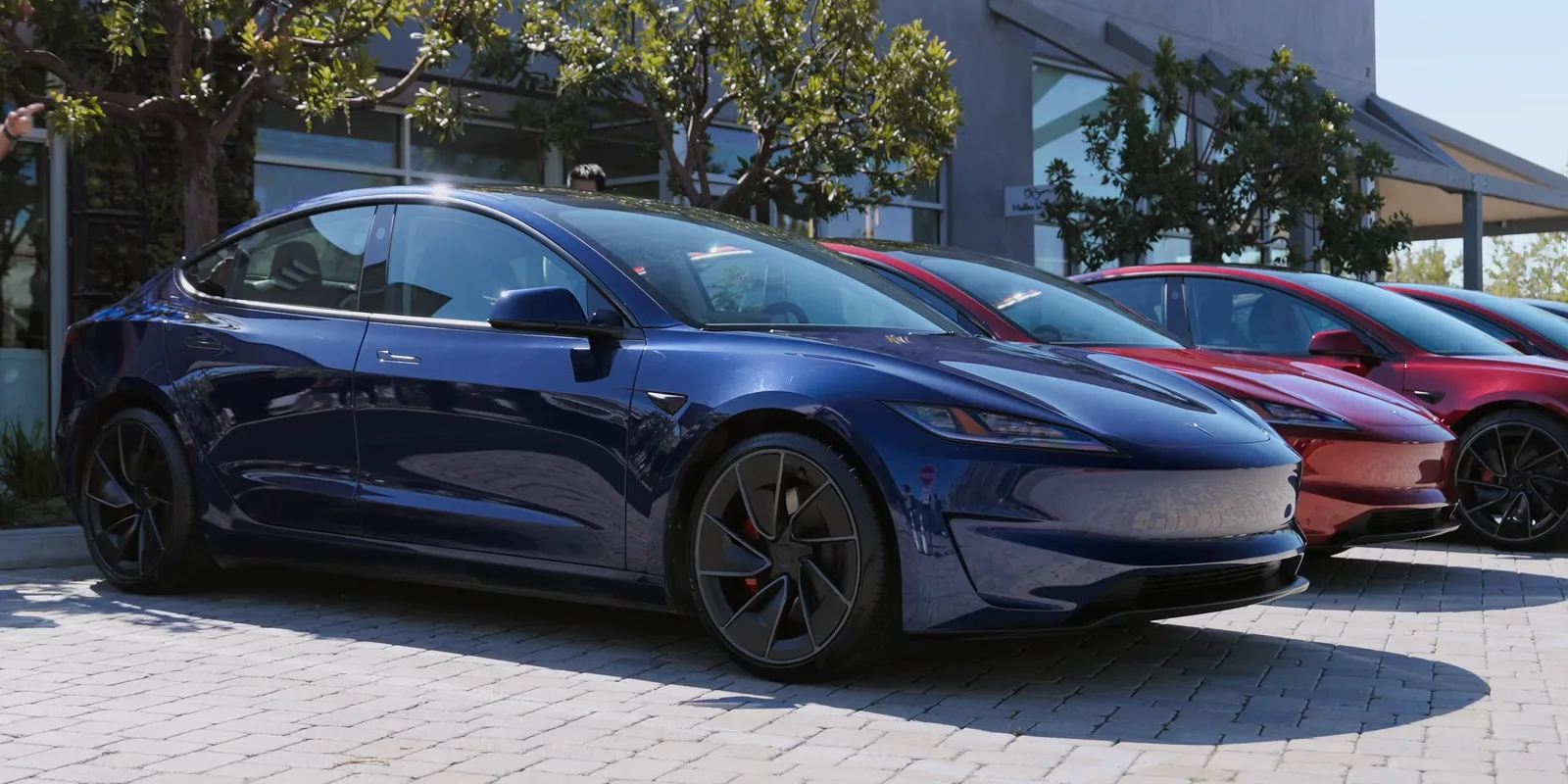 Mørkeblå Tesla Model 3 Performance set skråt forfra på brosten foran en Tesla-butik, med to røde Model 3 parkeret ved siden af og glasfacade og træer i baggrunden