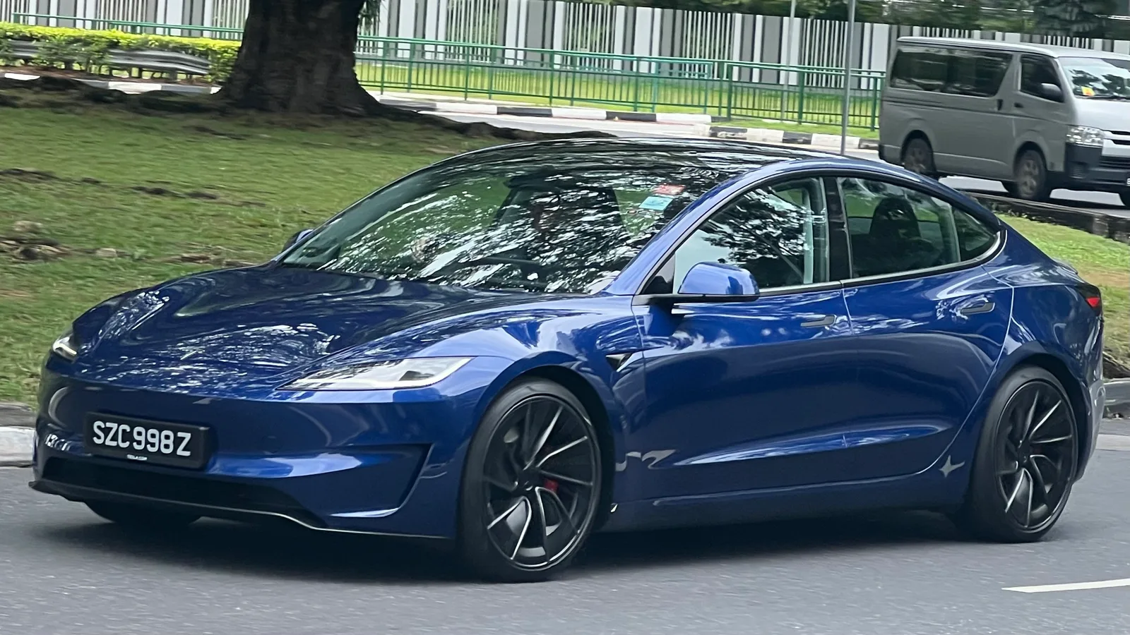 Mørkeblå Tesla Model 3 Performance set skråt forfra under kørsel på en vej langs en græsplæne, med røde bremsekalibre og sportsfælge