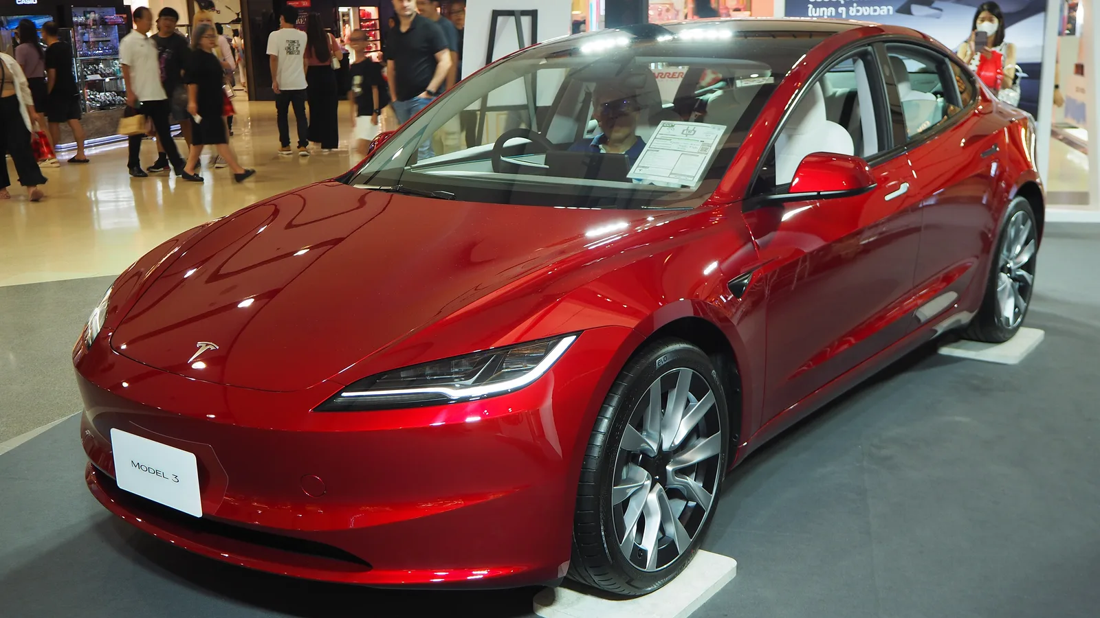 Rød Tesla Model 3 af facelift-udgaven set skråt forfra på en udstillingspodie, med den smalle lygtesignatur og et MODEL 3-skilt i nummerpladefeltet