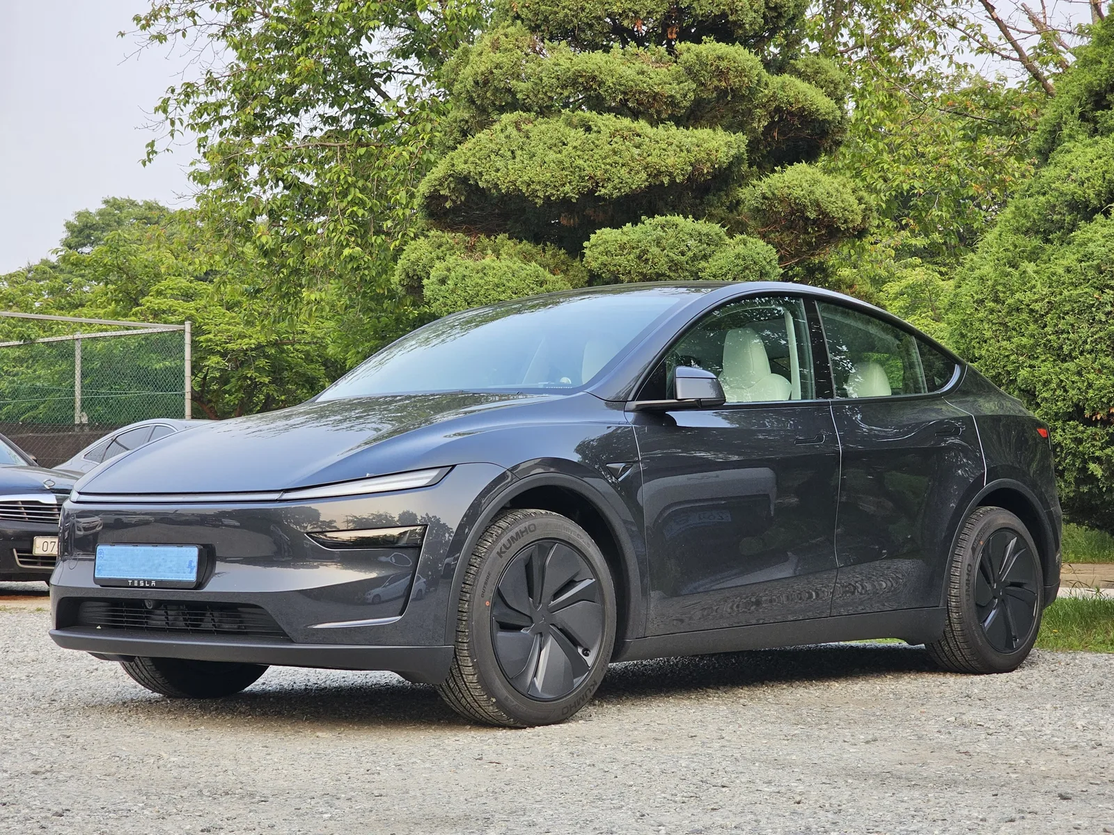 Grå Tesla Model Y af den nye udgave set skråt forfra, parkeret på grus med træer bag