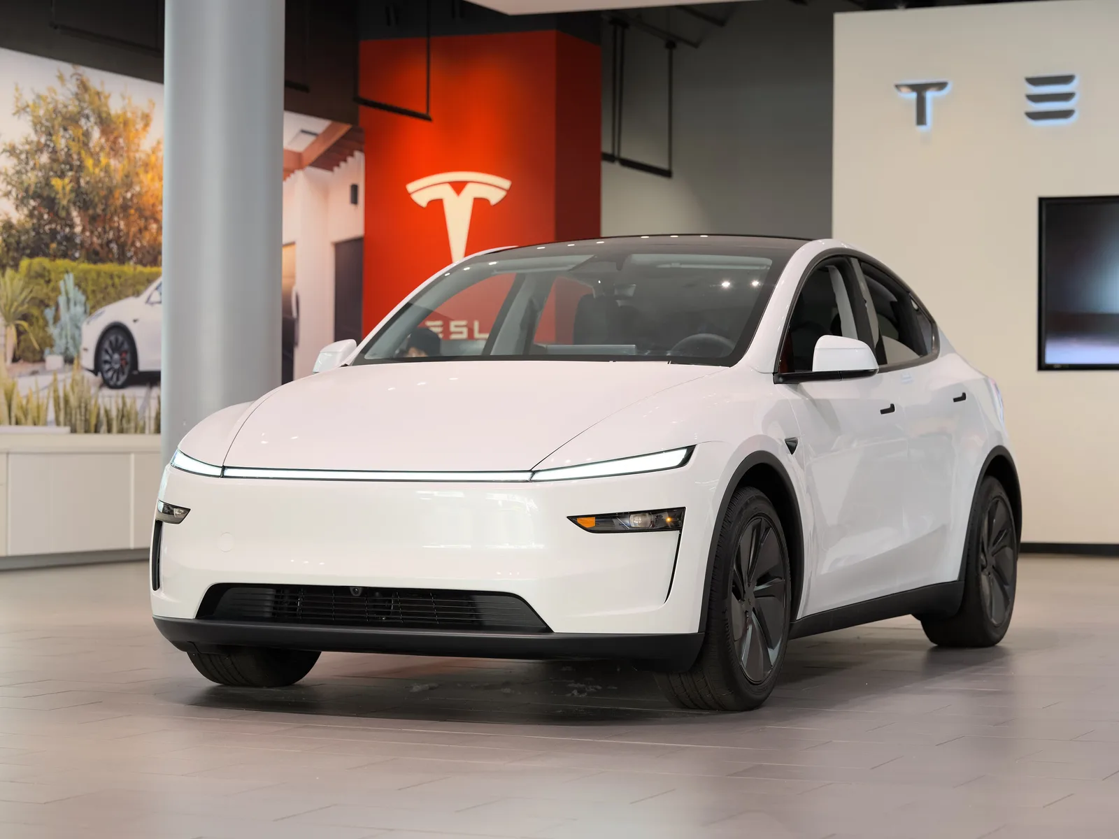 Hvid Tesla Model Y af 2025-udgaven set skråt forfra i en Tesla-butik, med en gennemgående lysstribe over et helt lukket næseparti