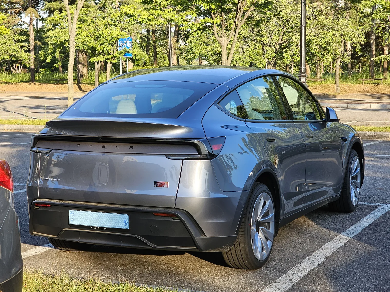 Grå Tesla Model Y af 2025-udgaven set skråt bagfra på en parkeringsplads ved en park i aftensol, med lyst interiør gennem ruderne
