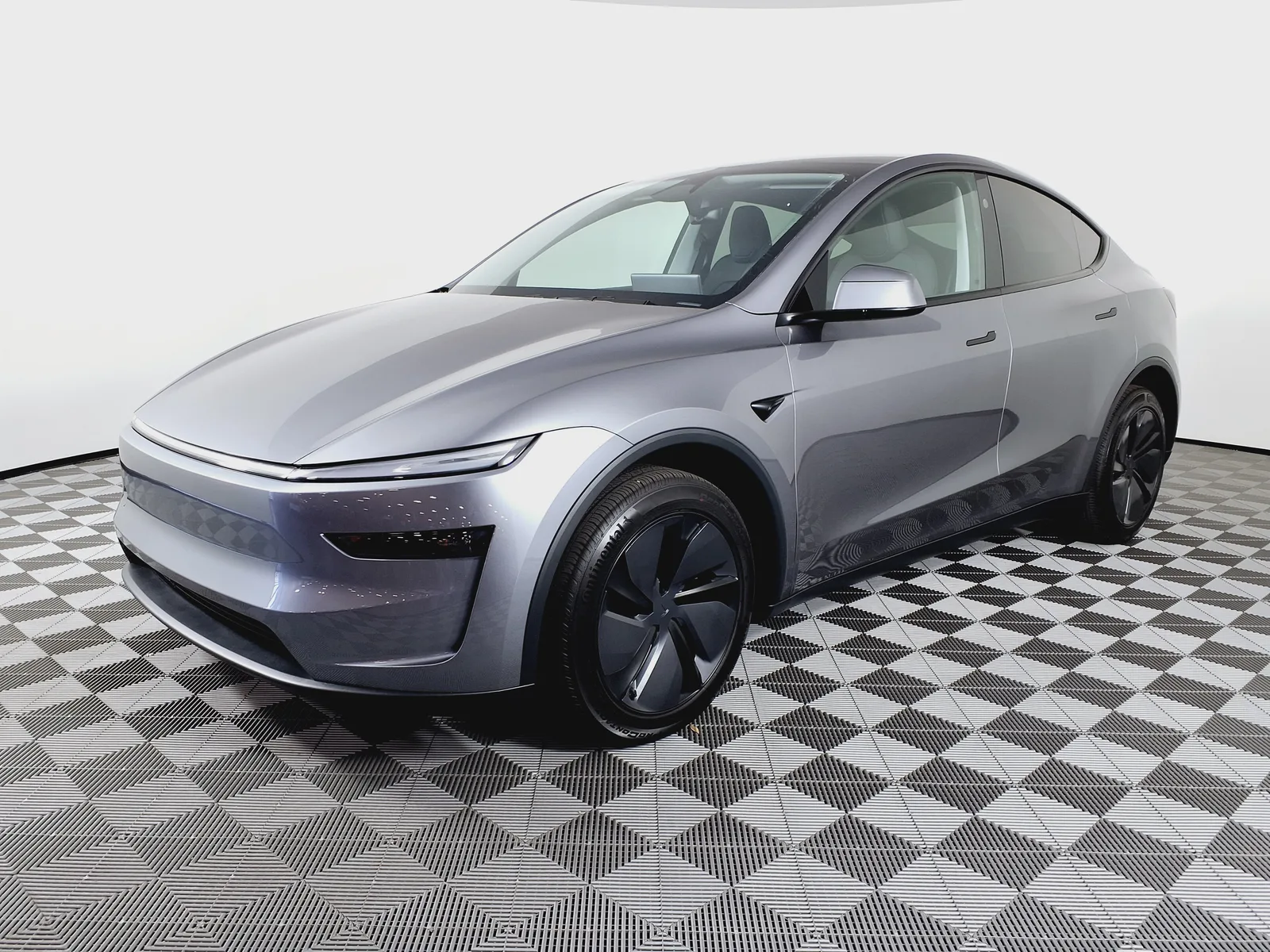 Grå Tesla Model Y af den nye udgave set skråt forfra i et fotostudie på ternet gulv, med lysbjælken tværs over fronten og aerofælge