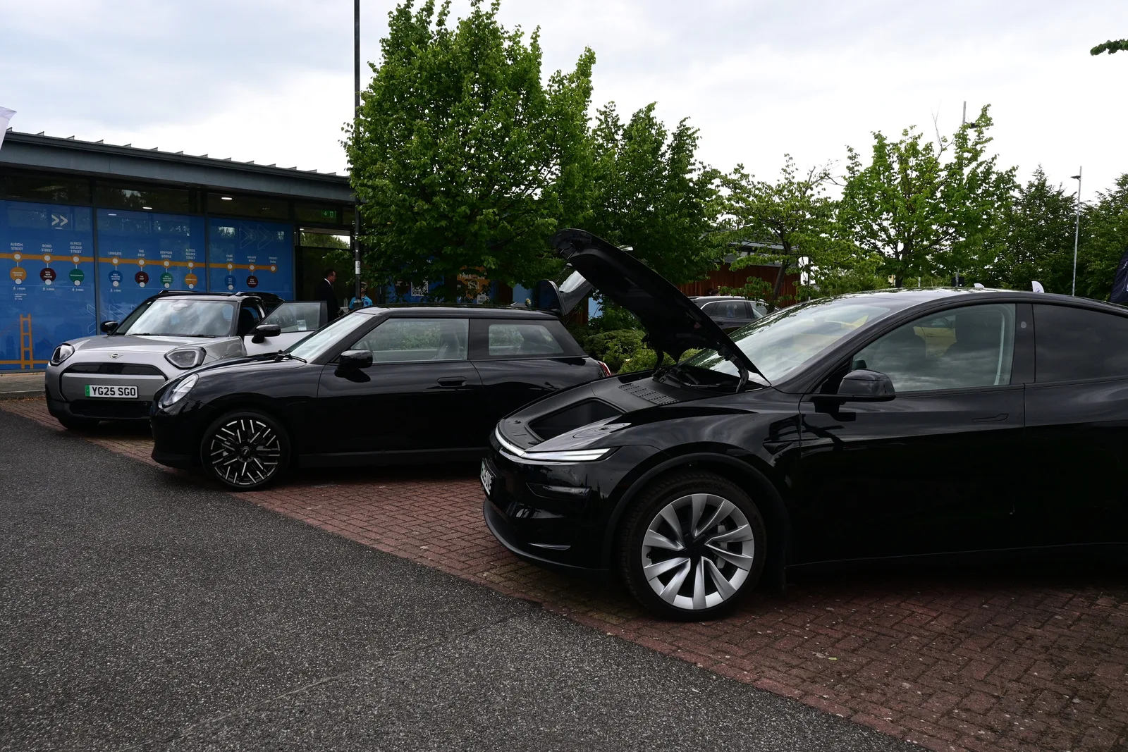 Sort Tesla Model Y af 2025-udgaven med frunken åben på en brostensbelagt plads, to Mini Cooper parkeret ved siden af.
