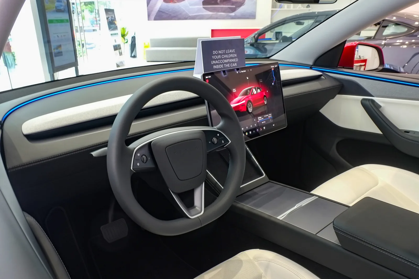 Førerpladsen i en ny Tesla Model Y set fra passagersiden, med lyst interiør, rat uden stilke og skærmen tændt