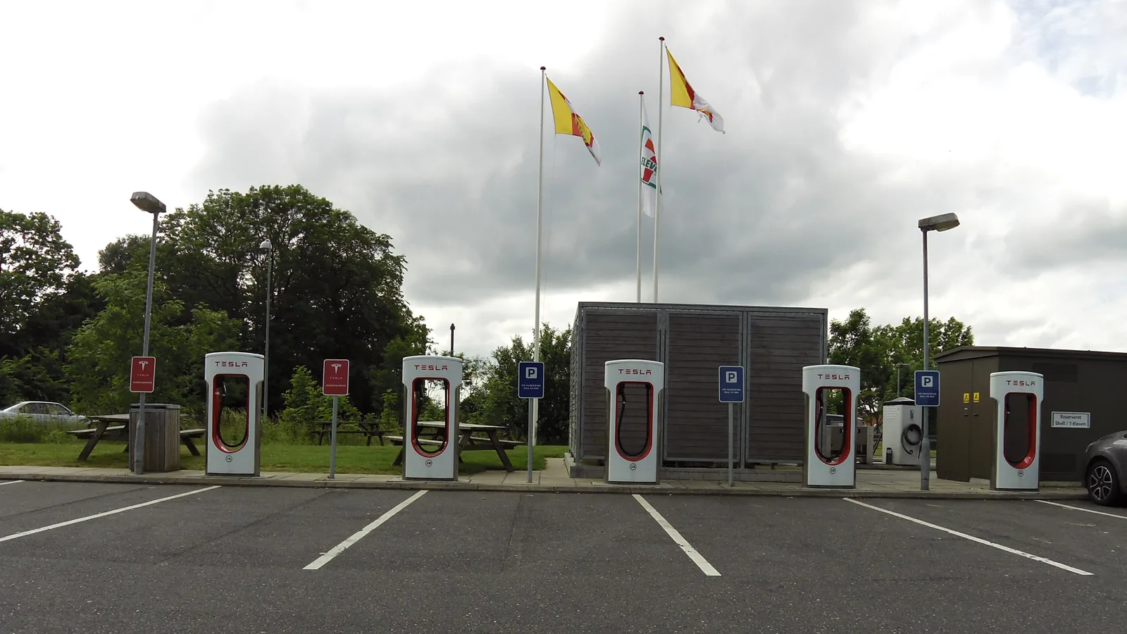 Fem hvide Tesla Superchargere i en række langs en tom parkeringsplads på en dansk rasteplads under en overskyet himmel