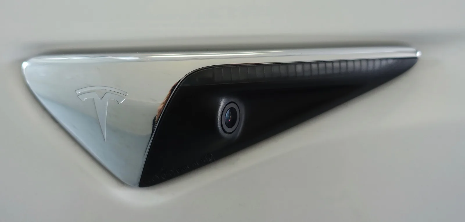 Nærbillede af sidekameraet i forskærmen på en hvid Model 3, i det sorte felt bag Tesla-mærket