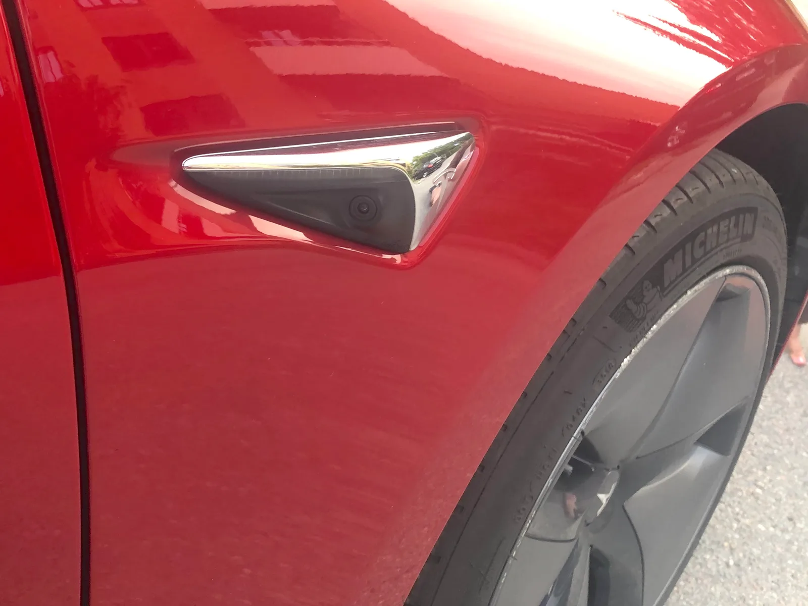 Nærbillede af den sorte sidekameraenhed i forskærmen på en rød Tesla Model 3, med hjulet og dækket bagved