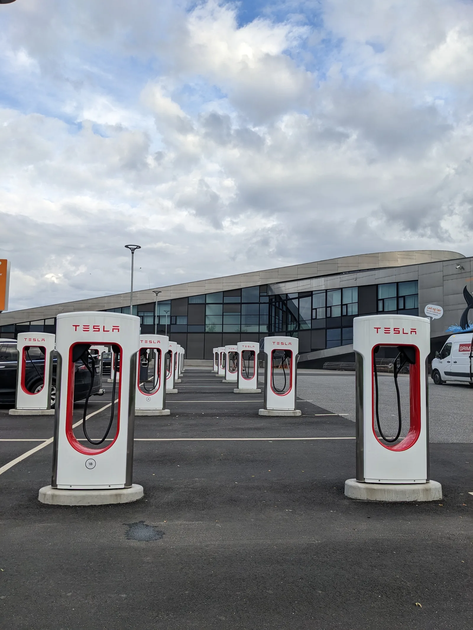 Et stort Supercharger-anlæg med en lang dobbeltrække af hvide standere på runde betonfundamenter, fotograferet ned gennem midtergangen foran en moderne bygning i glas og metal
