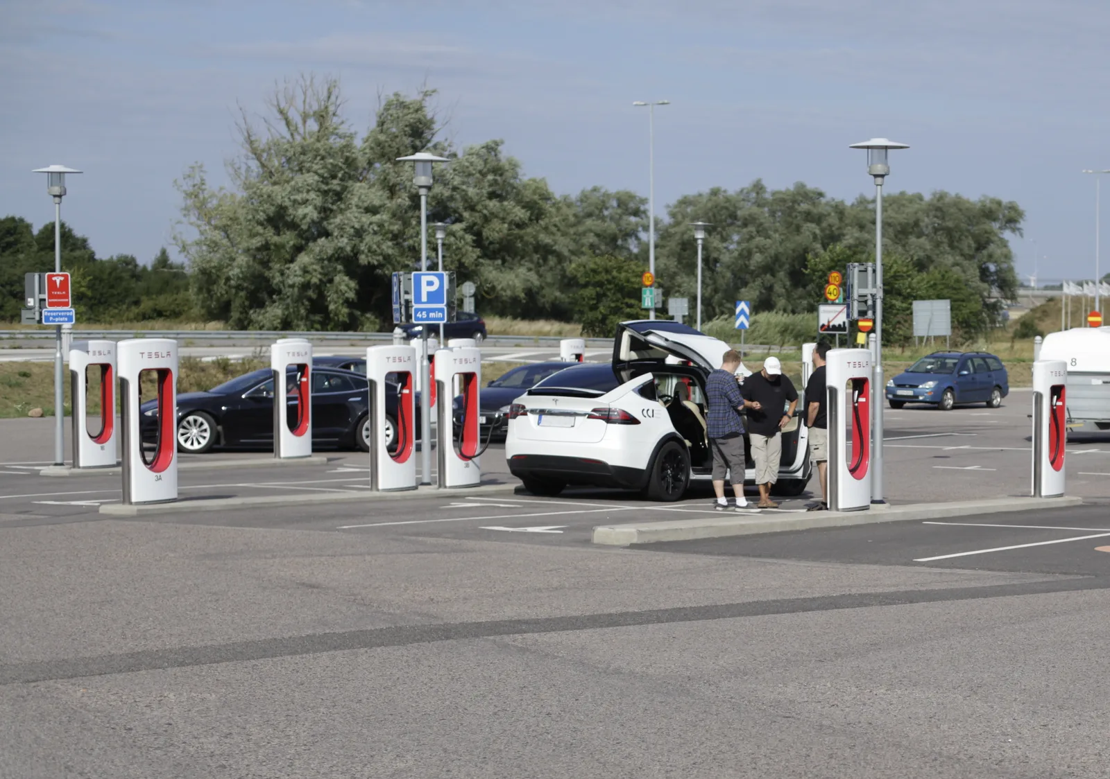 En hvid Tesla Model X med åbne falkevingedøre ved et Supercharger-anlæg, tre personer står ved bilen, og en sort Model S lader ved standeren bagved