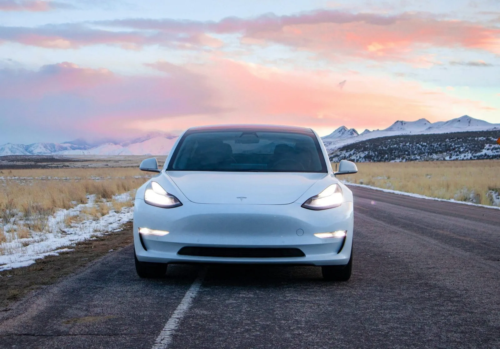 Hvid Tesla Model 3 af første generation set lige forfra med tændte lygter på en vej gennem et sneklædt landskab med bjerge i baggrunden