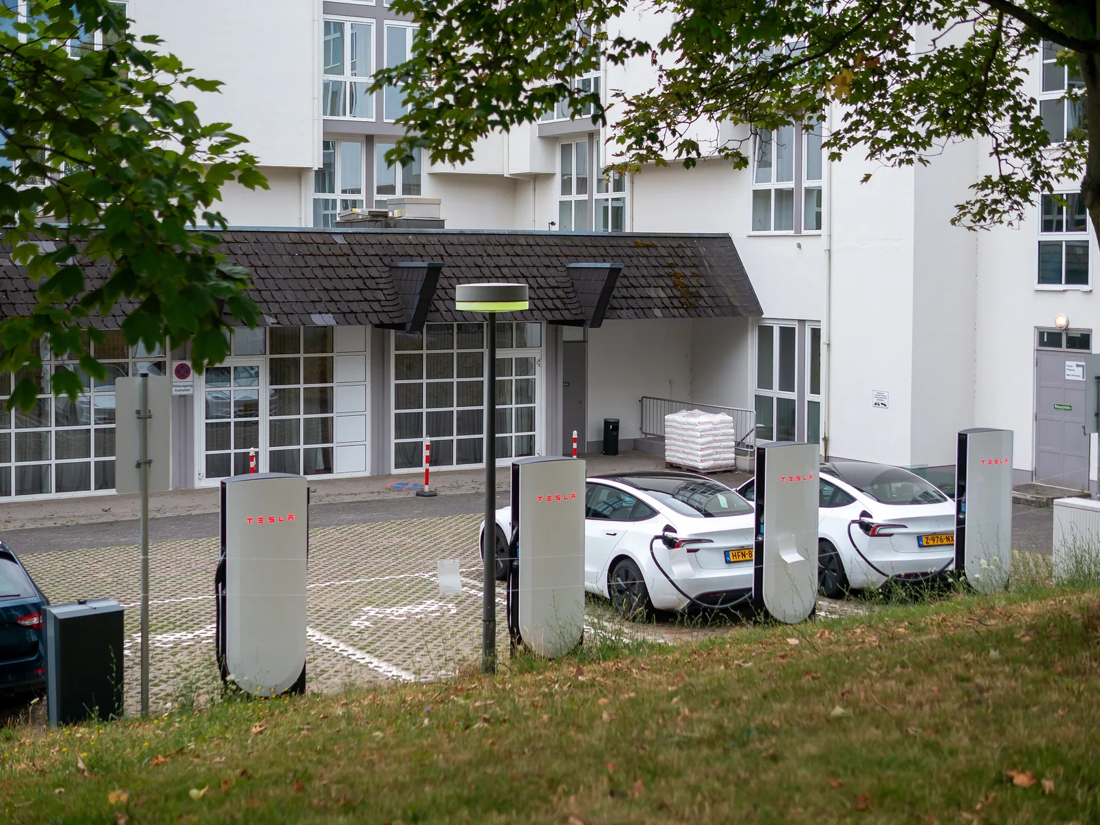 To slanke sorte V4-Superchargere med to hvide Tesla Model 3 tilsluttet på en brostensbelagt parkeringsplads foran en hvid bygning