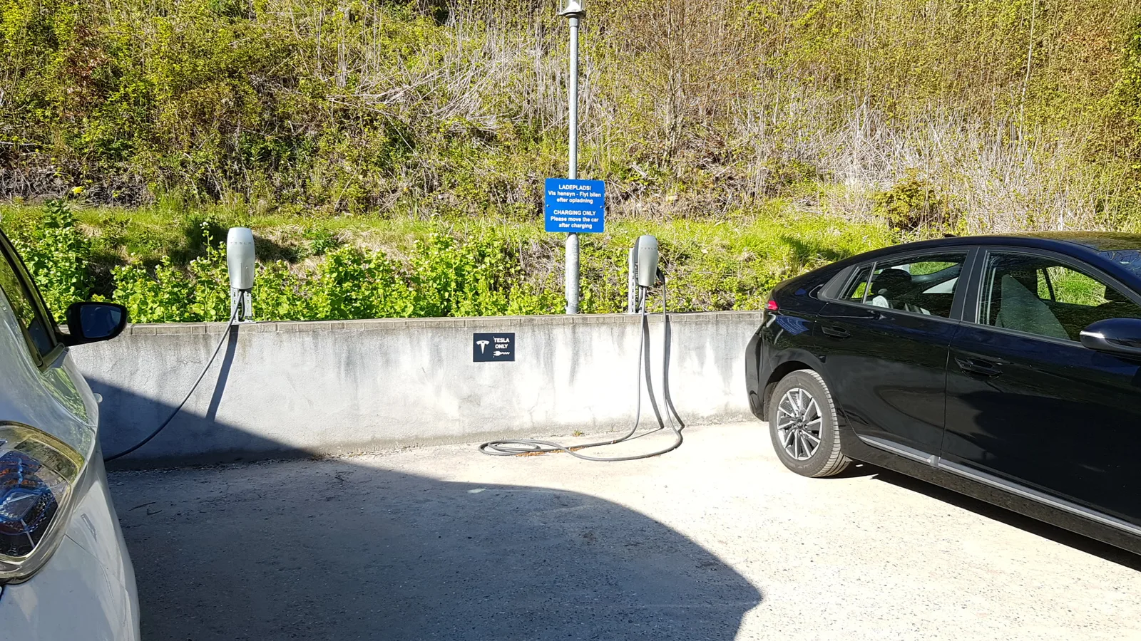 To Tesla Wall Connectors på en lav betonmur på en dansk hotelparkering med skiltet Ladeplads og to biler ved pladserne